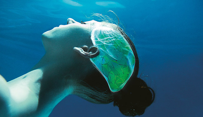 Eine Frau treibt mit geschlossenen Augen unter Wasser. Es sind nur ihr Hals und Kopf abgebildet. Auf der Seite ihres Kopfes befindet sich eine Grafik in Form eines Gehirns. Der hintere Teil der Grafik ist grün , wohingegen der vordere Teil in Türkis gefärbt ist.