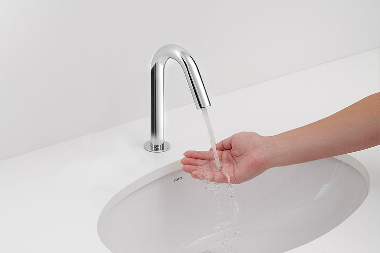 Das Bild zeigt eine Hand unter fließendem Wasser eines modernen, chromglänzenden Wasserhahns von TOTO, der über einem Waschbecken installiert ist.