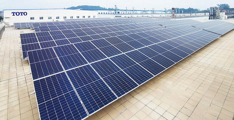 Das Foto zeigt eine große Anordnung von Solarpaneelen auf dem Dach eines Industriegebäudes mit dem Logo von TOTO im Hintergrund.