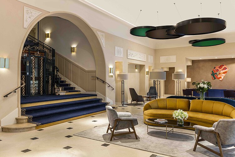 Das Bild zeigt das Innere einer eleganten und geräumigen Hotellobby mit einer Mischung aus klassischer und moderner Einrichtung. Im Zentrum steht ein großes, gelbes, halbkreisförmiges Sofa, flankiert von zwei grauen Sesseln, während im Hintergrund eine geschwungene Treppe mit blauem Teppich und schmiedeeisernem Geländer zu sehen ist.