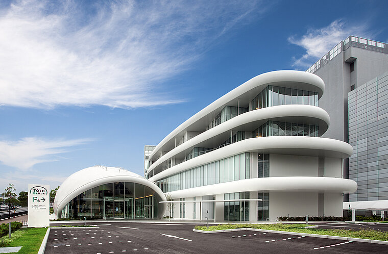 Das Bild zeigt ein modernes Gebäude mit einer dynamischen, geschwungenen weißen Fassade und großen Fenstern unter einem blauen Himmel. Vor dem Gebäude befindet sich ein Schild mit der Aufschrift "TOTO".