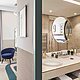 L'image est un collage de deux pièces : sur la gauche, une partie d'une chambre à coucher avec un siège bleu et une table d'appoint ronde près d'une fenêtre, tandis que sur la droite, une salle de bain moderne avec deux lavabos, un grand miroir rond et une étagère éclairée. Les pièces sont décorées dans un style élégant et contemporain, avec des couleurs neutres et des accents de bleu et d'or.