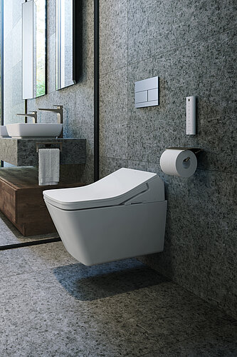 Das Bild zeigt einen Teil eines modernen Badezimmers mit einer wandhängenden Toilette mit geschwungenem Design vor einer grauen Wand mit strukturierter Fliese. Zu sehen ist auch ein Teil der Holzeitelkeit mit einem darauf befindlichen Handtuch und einem Spiegel, der die Szene reflektiert.