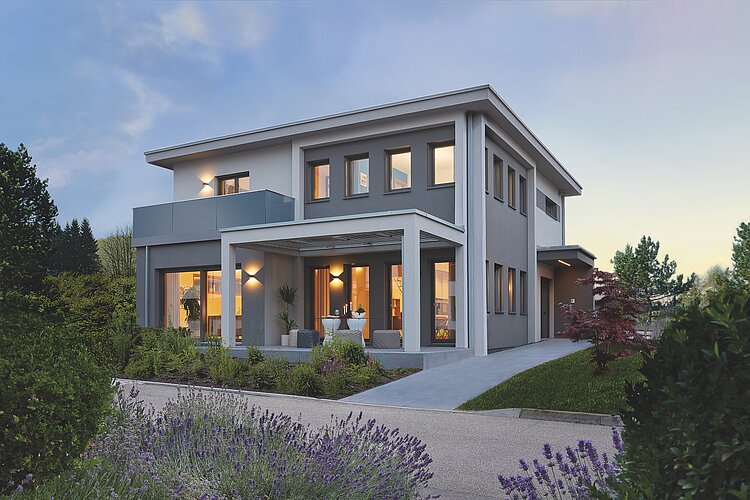 La photo montre une maison moderne de deux étages au crépuscule, dont l'éclairage intérieur crée une ambiance accueillante. La maison est entourée d'un jardin paysager bien entretenu, avec de la lavande en fleurs au premier plan, et dispose d'une terrasse spacieuse avec des sièges.