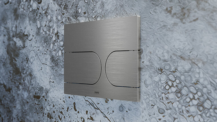 Das Bild zeigt eine moderne, gebürstete Metall-Druckplatte für eine Toilette mit zwei Tasten, montiert auf einer Wand mit einer auffälligen Marmoroptik. Die Platte trägt das Logo von TOTO und hebt sich durch ihr schlichtes Design deutlich von der lebhaft gemusterten Wand im Hintergrund ab.
