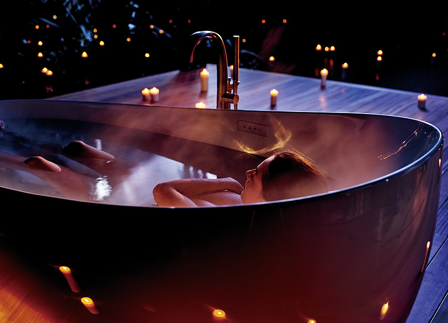 A woman lies in an oval bathtub. Everything except her knees, head and arms crossed over her chest is covered by water. There are candles in the background.