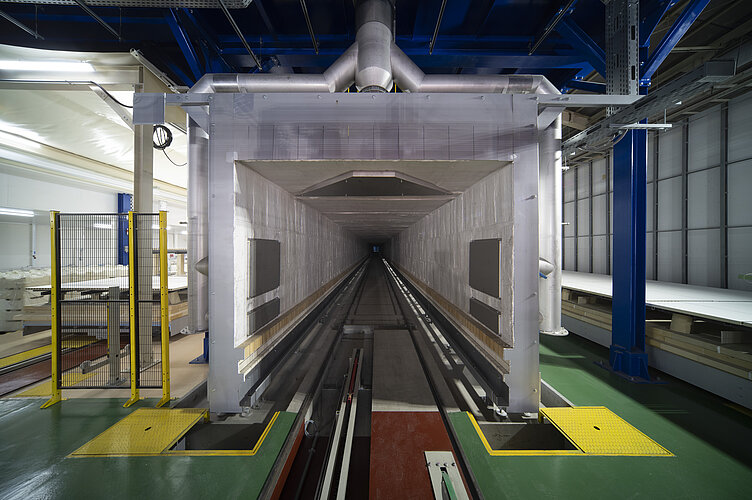 Das Bild zeigt das Innere einer industriellen Anlage mit einem großen, tunnelartigen Ofen, der durch Schienen im Boden für den Transport von Materialien ausgelegt ist. Über dem Eingang des Tunnels befindet sich eine große Abluftanlage, und die Umgebung ist mit Sicherheitsgeländern und Warnfarben für Sicherheitszonen gekennzeichnet.