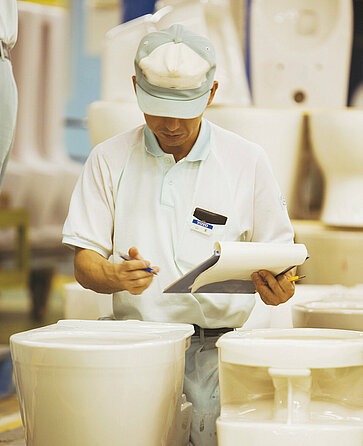 A man stands in front of some WC ceramics and looks at them closely. He is holding a clipboard with paper and a pen in his hands.