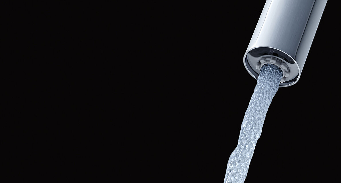 Close-up of the outlet of a faucet with ECOCAP against a black background. The water jet coming out of the tap is interspersed with small air bubbles. 