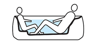 Two people sit in a bath with recline comfort technology. The taller person sits on the right-hand side of the bath, the shorter person on the left. She sits on a small elevation in the base of the bath. There is enough space for both to sit comfortably. 