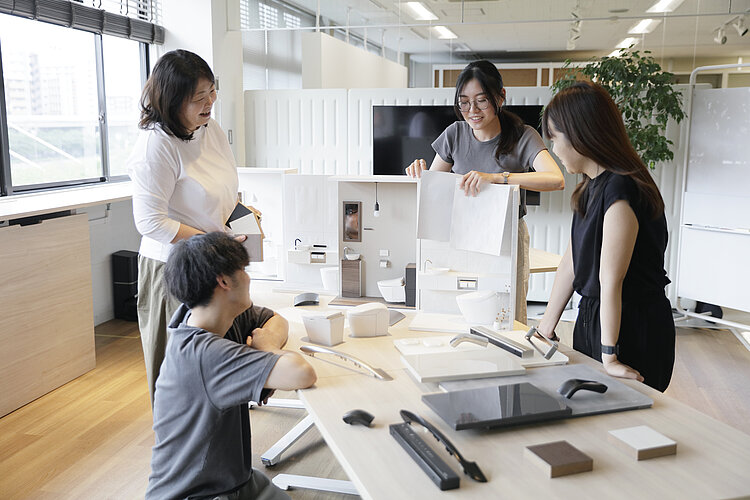 Das Bild zeigt eine Gruppe von vier Personen, die in einem hellen Büro um einen Tisch mit verschiedenen Materialmustern und Modellen für Badezimmerdesign stehen. Zwei Frauen stehen und scheinen ein Gespräch zu führen, während ein Mann sitzt und einem weiteren Gegenstand auf dem Tisch Beachtung schenkt, und eine weitere Frau, ebenfalls stehend, ein Blatt Papier hält.
