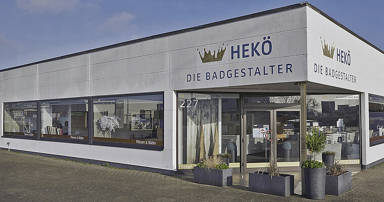 Auf dem Bild ist das Gebäude eines Fachgeschäfts mit dem Namen "HEKÖ DIE BADGESTALTER" zu sehen, welches auf Badezimmerdesign und -renovierung spezialisiert ist. Das eingeschossige Eckgebäude hat eine große Fensterfront, durch die Ausstellungsstücke sichtbar sind, und ist mit Pflanzenkübeln vor dem Eingang dekoriert.