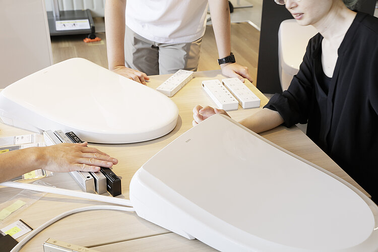 Auf dem Foto sind die Hände von drei Personen zu sehen, die um einen Tisch herumstehen, auf dem zwei abgenommene Toilettensitze und mehrere Fernbedienungen für Toilettensysteme liegen. Die Personen scheinen verschiedene Modelle oder Funktionen der Toilettentechnologie zu prüfen oder zu vergleichen, wobei eine Person in Schwarz gekleidet ist, die eine der Fernbedienungen berührt.