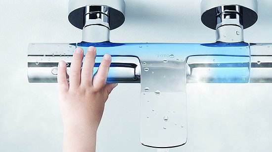 Ein Kind fasst an eine Duscharmatur, mit zwei Zuläufen. Der Bereich um den Ausfluss und die Zuläufe ist blau gekennzeichnet. 