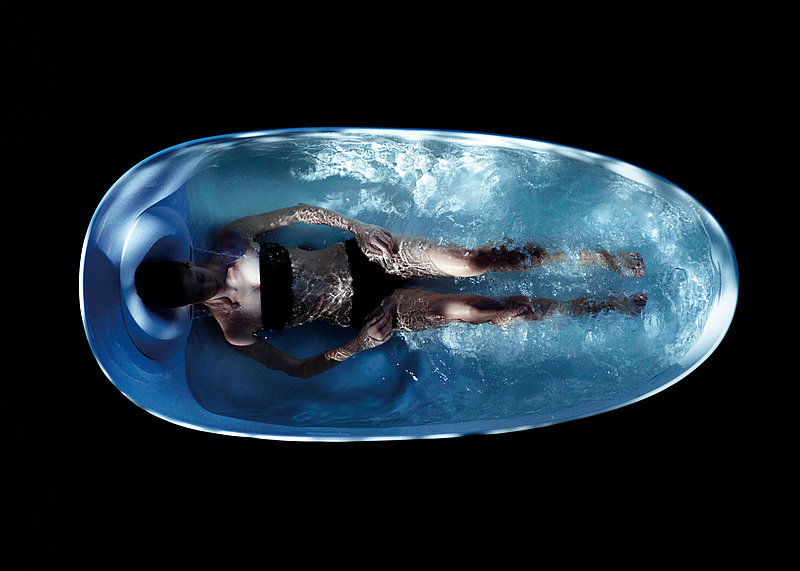 A bird's eye view of a woman lying in a floating bathtub. The background is black.