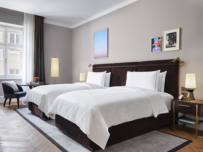 Das Bild zeigt ein ansprechend eingerichtetes Schlafzimmer mit zwei nebeneinanderstehenden Betten mit dunklen Kopfteilen und weißer Bettwäsche. An den Wänden hängen moderne Kunstwerke, und das Zimmer wird durch Tageslicht von einem großen Fenster und warmes Licht von Tischlampen beleuchtet, was eine gemütliche Atmosphäre schafft.