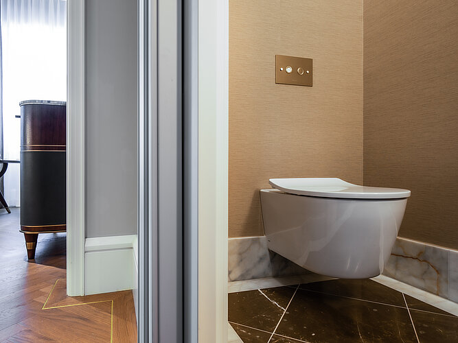 Das Foto zeigt eine Ansicht einer modernen, wandhängenden Toilette in einem eleganten Badezimmer mit beige gefärbten Wänden und einem dunkel gemusterten Marmorfußboden. Links im Bild ist teilweise ein luxuriöses, dunkles Holzmöbel zu sehen, und an der Wand über der Toilette befindet sich eine stilvolle Bedienplatte für die Spülung.