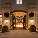 Das Bild zeigt den festlich beleuchteten Eingangsbereich eines eleganten Hotels, geschmückt mit Kränzen und Lichterketten. Laternen und mit Lichtern dekorierte Pflanzenkübel flankieren den Weg, der zu den automatischen Glastüren führt, und verleihen dem Eingang eine einladende und festliche Atmosphäre.