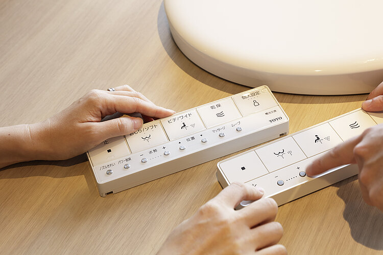 Das Foto zeigt die Hände von zwei Personen, die Fernbedienungen für ein Badezimmersystem bedienen, auf denen in japanischer Sprache verschiedene Funktionen und Einstellungen beschriftet sind. Eine Hand drückt auf einen Knopf, während die andere Hand eine andere Fernbedienung hält, die zu einem Produkt der Marke TOTO gehört, was auf einen Kontext der Produktprüfung oder -demonstration hindeutet.