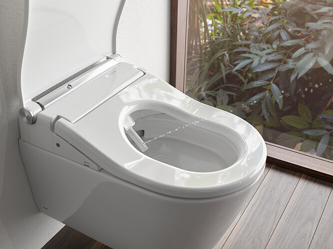 Das Foto zeigt eine Nahaufnahme eines modernen, weißen Toilettenbeckens mit geöffnetem Deckel und integrierter Bidetfunktion, sichtbar durch die im Sitz eingelassene Düse. Die Toilette befindet sich neben einem Fenster, durch das man auf grüne Pflanzen blicken kann, was auf ein Badezimmer mit natürlichem Licht und Blick nach draußen hindeutet.