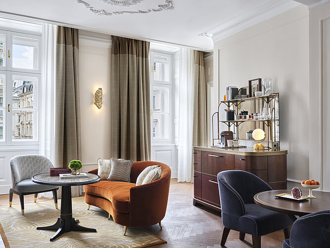 Das Bild zeigt ein stilvoll eingerichtetes Wohnzimmer mit klassischen Stuckdecken und großen Fenstern, die Tageslicht hereinlassen. In dem Zimmer stehen ein orangefarbener Sessel, zwei blaue Stühle um einen runden Tisch, und es gibt eine elegante Holzkonsole mit einem Spiegel und dekorativen Objekten darauf.