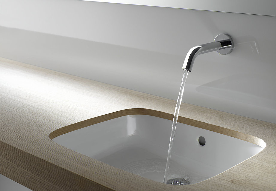 A tap is mounted on a wall above a washbasin. The tap is switched on and a jet of water is coming out of it. 