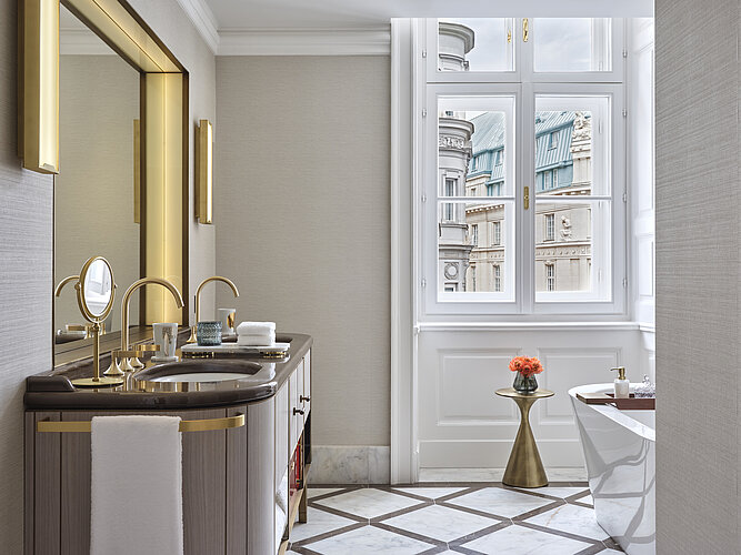 Das Bild zeigt ein luxuriöses Badezimmer mit einem großen Fenster, das einen Blick auf historische Gebäudefassaden bietet. Im Vordergrund ist eine elegante Waschtischanlage mit goldenen Armaturen zu sehen, und rechts im Bild erkennt man den Rand einer freistehenden Badewanne.