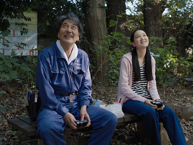Hirayama und seine Nichte Niko sitzen auf eine Bank und schauen lächelnd nach oben.