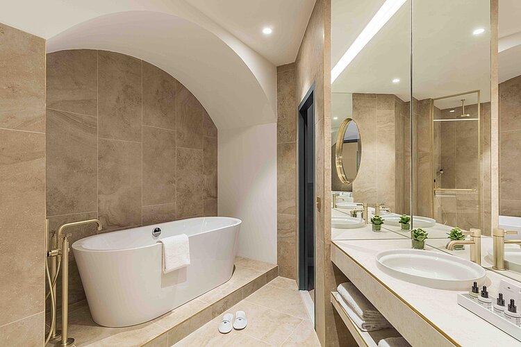 The picture shows a luxurious bathroom with neutral tones and modern fittings. A free-standing bathtub is located in an alcove with a vaulted ceiling, while a long washbasin with two washbasins, large mirrors and gold-colored fittings can be seen opposite.