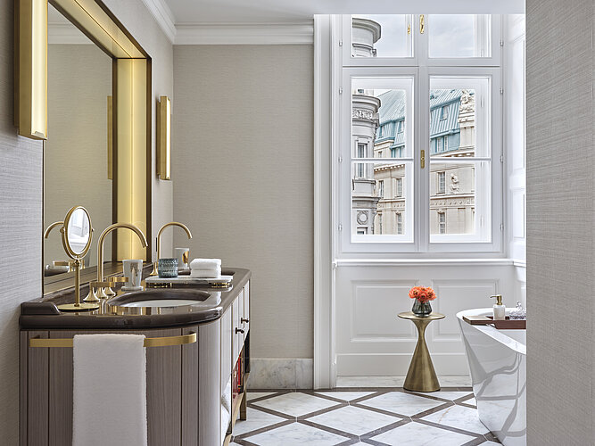 L'image montre une salle de bains luxueuse avec une grande fenêtre qui offre une vue sur les façades de bâtiments historiques. Au premier plan, on peut voir un élégant lavabo avec une robinetterie dorée, et à droite de l'image, on distingue le bord d'une baignoire sur pied.