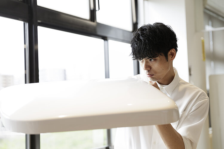 The picture shows a young man with black hair looking intently at the design of a toilet lid in the lower part of the picture, which he is holding with both hands. He is wearing a white shirt and is standing in a brightly lit room with large windows in the background.