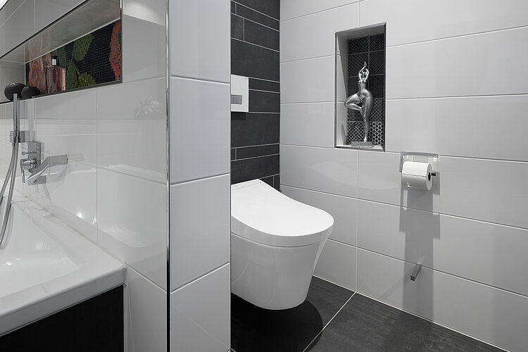 Das Foto zeigt ein Teil eines Badezimmers mit einem modernen Washlet (eine Kombination aus Toilette und Bidet) neben einer Badewanne, die beide von weiß und dunkelgrau gefliesten Wänden umgeben sind. Auf der gegenüberliegenden Wand ist ein kleines Fenster mit einem Gitter, hinter dem eine dekorative Figur zu sehen ist.