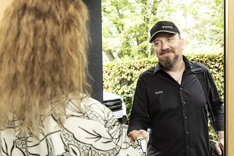 Das Bild zeigt einen Mann mit einer schwarzen Kappe und einem schwarzen Hemd mit dem Logo "TOTO" darauf, der freundlich lächelt und einer Frau mit dem Rücken zur Kamera die Hand schüttelt. Im Hintergrund ist ein grüner Garten zu erkennen, was darauf hindeutet, dass die Szene vor einem Wohnhaus spielt.