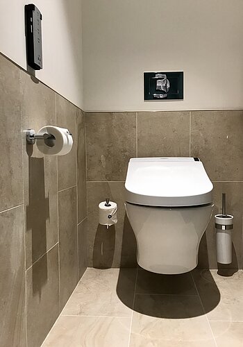 The photo shows a simple and clean toilet cubicle with modern elements. Tiled beige tones on the wall surround a white WASHLET, next to it is a toilet roll holder, a small waste garbage can and a brush, while a dispenser for hygiene products is mounted above it.