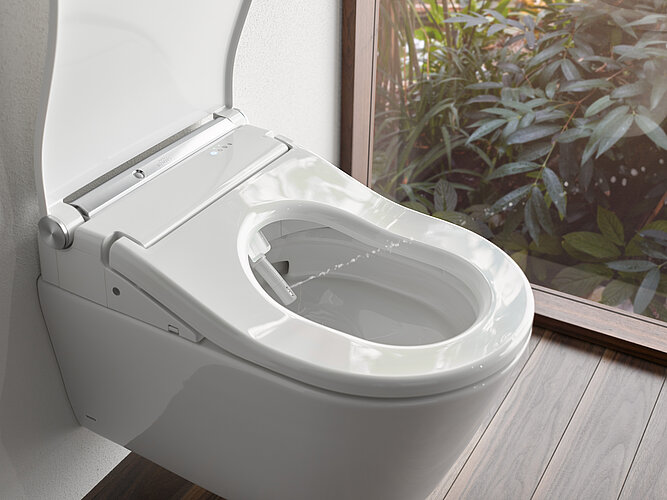 The picture shows a close-up of a modern, wall-hung TOTO WASHLET with an open seat cover and integrated bidet function, visible through the nozzle embedded in the seat. The toilet stands next to a window with a view of green plants, suggesting a bathroom with natural light and a view of the outside.
