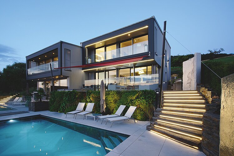 The picture shows a modern, cubic house with two floors at dusk, highlighted by the interior and exterior lighting. An illuminated staircase leads up to the house and there is a swimming pool in front of the building, surrounded by sun loungers and carefully landscaped gardens.