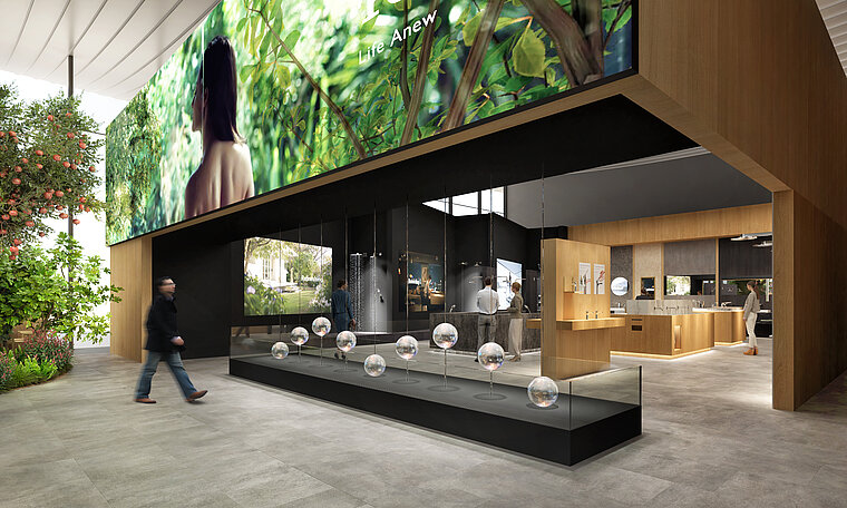 The image shows a modern exhibition hall with a dark-colored, open TOTO stand presenting shower and faucet products, accompanied by clear ball displays on a long, black counter. A large screen display with images of nature can be seen above the stand, while a green plant wall on the left emphasizes the natural and environmentally conscious orientation of the brand.