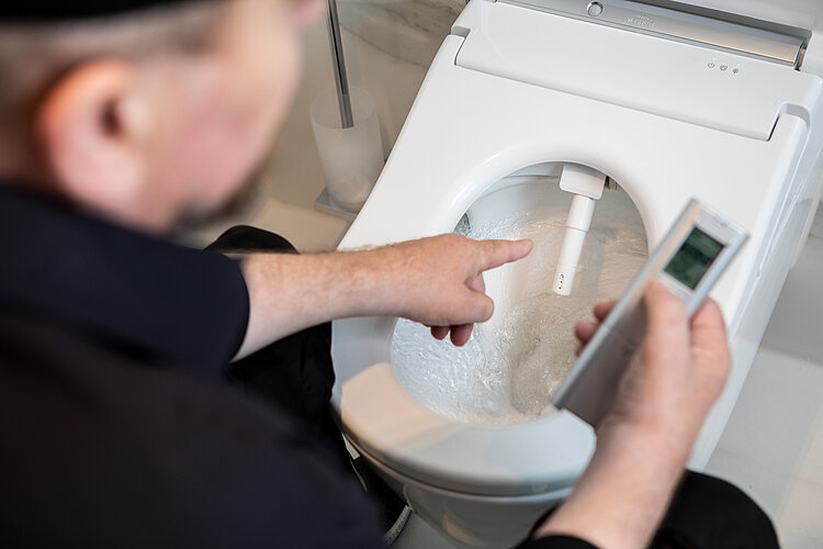 Das Foto zeigt eine Nahaufnahme eines Mannes, der eine schwarze Kappe trägt und auf ein Washlet deutet, während Wasser in die Toilette gespült wird. Er hält ein Steuergerät in der anderen Hand, möglicherweise um die Funktionen des Washlets zu demonstrieren oder zu testen.
