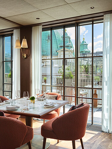 The picture shows an attractively decorated dining room with a round glass dining table and rust-colored chairs, illuminated by elegant pendant lights. The large windows offer a view of a terrace and the historic architecture in the background, suggesting an urban location.