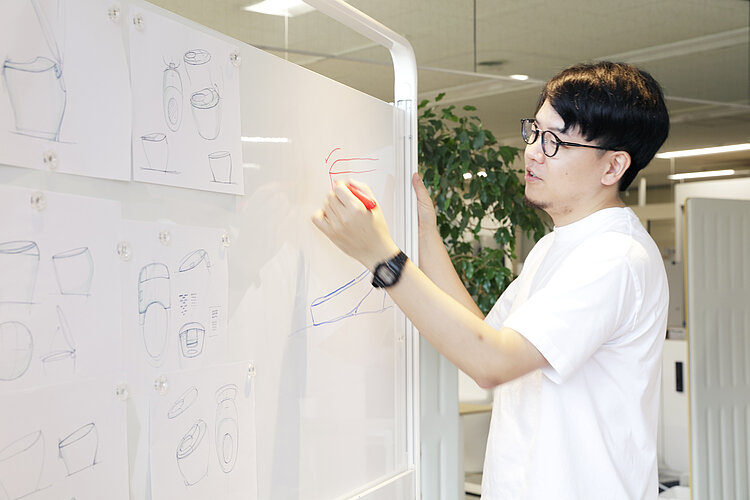 Das Bild zeigt einen Mann mit Brille und schwarzem Haar, der ein weißes Hemd trägt und an einer weißen Tafel mit verschiedenen Skizzen von Badezimmerartikeln steht. Er scheint mit einem roten Marker eine Skizze zu zeichnen oder zu annotieren, während er sich auf die dargestellten Konzepte konzentriert.