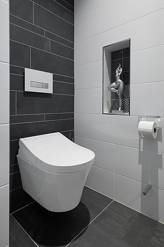 L'image montre une salle de bains moderne avec des toilettes suspendues à un mur carrelé. Au-dessus des toilettes se trouve une petite étagère en niche avec une figurine décorative, et la palette de couleurs de la pièce se compose de tons gris foncés et clairs.