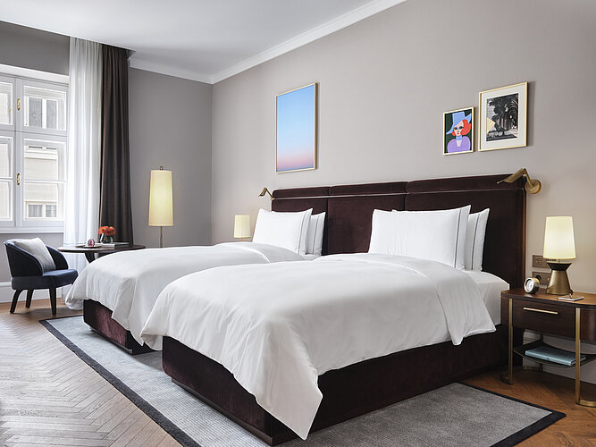 The picture shows an attractively furnished bedroom with two adjacent beds with dark headboards and white bed linen. Modern artwork hangs on the walls and the room is illuminated by natural light from a large window and warm light from table lamps, creating a cozy atmosphere.