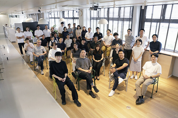 The picture shows a large group of people who have positioned themselves for a group photo in an office or workshop room with modern furnishings and large windows. They are dressed casually, some are sitting on chairs in the front rows while others are standing behind them, all looking towards the camera.