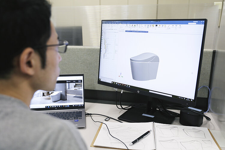 Das Bild zeigt einen Mann, der vor einem Computerbildschirm sitzt, auf dem eine 3D-Konstruktionszeichnung eines Badezimmerprodukts zu sehen ist. Auf dem Schreibtisch vor ihm liegen Skizzen und ein geöffnetes Notizbuch, daneben ein Laptop, der ein Bild von einem Badezimmer zeigt, was darauf hindeutet, dass er an einem Design- oder Ingenieursprojekt arbeitet.