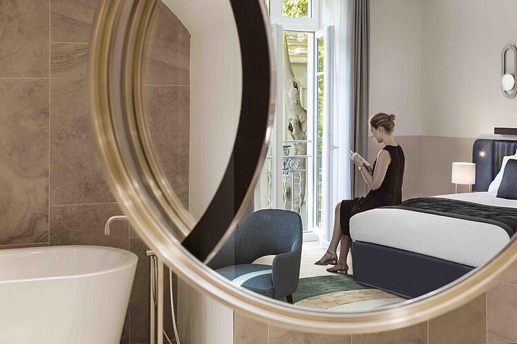 The photo shows a view through a round, wall-mounted mirror of a stylish suite with a modern interior. A woman in an elegant dress is visible through the mirror, sitting in an armchair on the balcony and reading a book, while a free-standing bathtub can be seen in the foreground of the mirror.