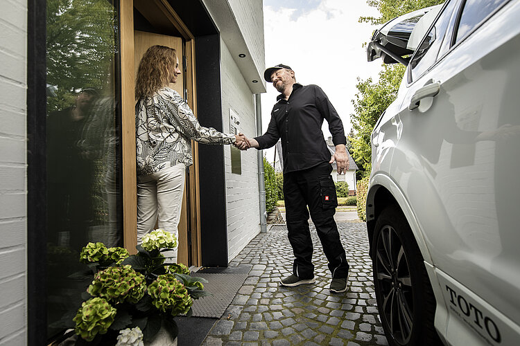Das Bild zeigt eine Szene vor einem Haus, wo eine Frau in einer hellen Bluse und Hose die Haustür öffnet und einem Mann in dunkler Kleidung die Hand schüttelt. Ein weißes Auto steht im Vordergrund, und die Umgebung lässt auf eine ruhige Wohngegend schließen.