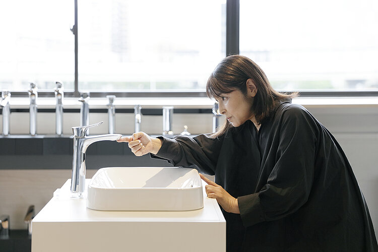 The photograph shows a woman wearing a black top and bending over a white sink in a bright room to check or operate the faucet. In the background are large windows letting in daylight and a row of identical taps, suggesting that the scene is in an exhibition space.
