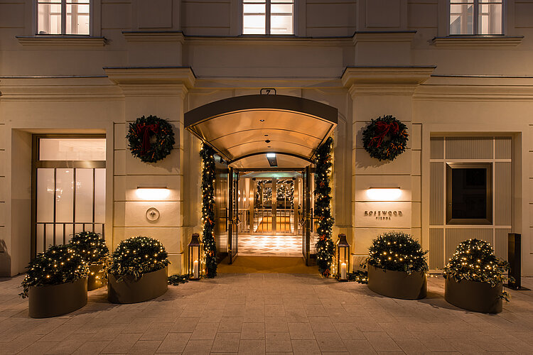 The picture shows the festively illuminated entrance area of an elegant hotel, decorated with wreaths and fairy lights. Lanterns and planters decorated with lights flank the path leading to the automatic glass doors, giving the entrance an inviting and festive atmosphere.