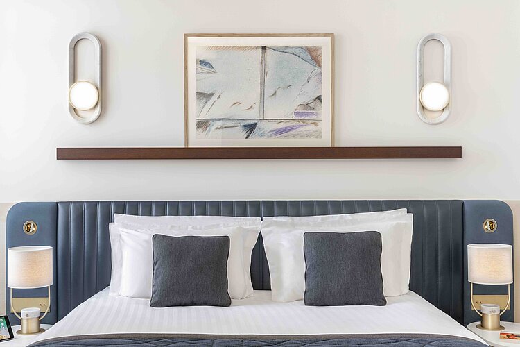 The picture shows the top of a bed in a brightly decorated room, with a dark blue, tufted headboard furnished with several cushions in gray and white. A framed work of art hangs above the bed, flanked by two modern, oval wall lamps that emit a warm light.