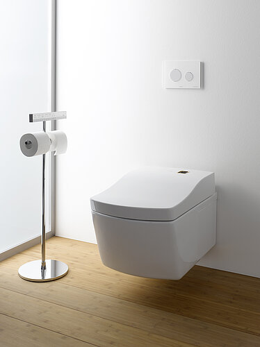 Das Bild zeigt eine minimalistische, wandhängende Toilette mit geschlossenem Deckel neben einer Toilettenpapierhalterung auf einem Holzfußboden, mit einer weißen Wand im Hintergrund. Über der Toilette befindet sich eine moderne, weiße Bedienplatte für die Spülung, die das schlichte und moderne Design des Raumes unterstreicht.